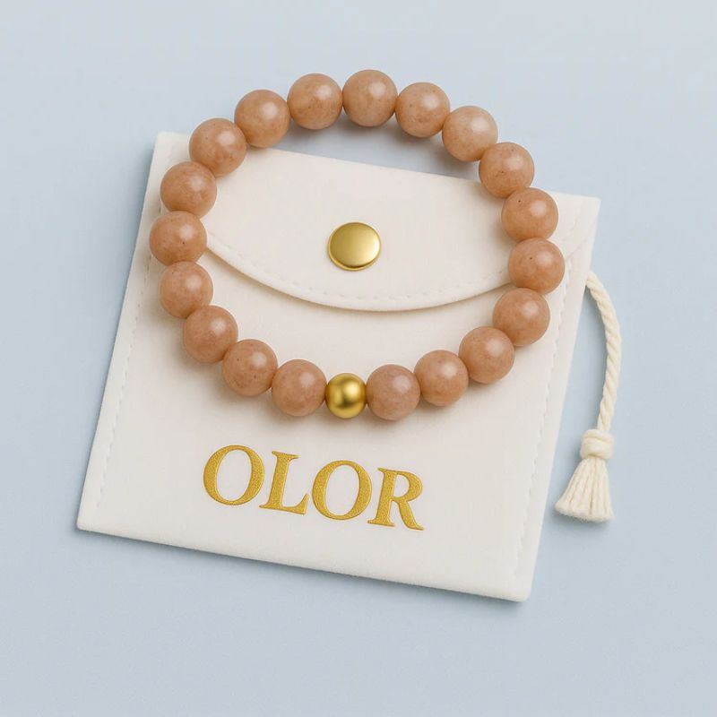 Sonnenstein bracelet with 6mm round beads on elastic band, warm orange gemstone displayed on white OLOR pouch with gold button and tassel.