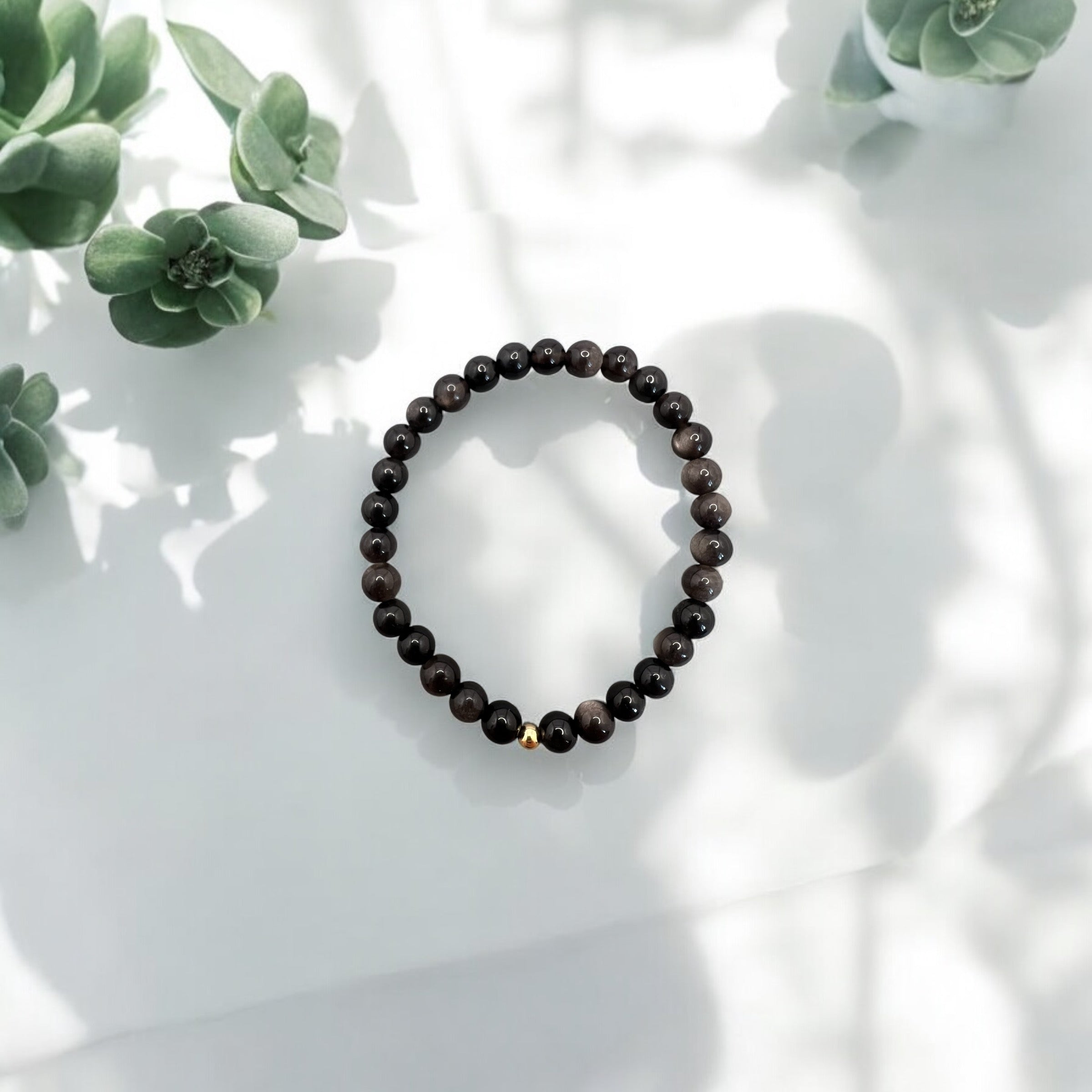 Goldobsidian Armband mit schwarzen Perlen und goldener Verschlussperle auf weißem Hintergrund mit grünen Pflanzenakzenten