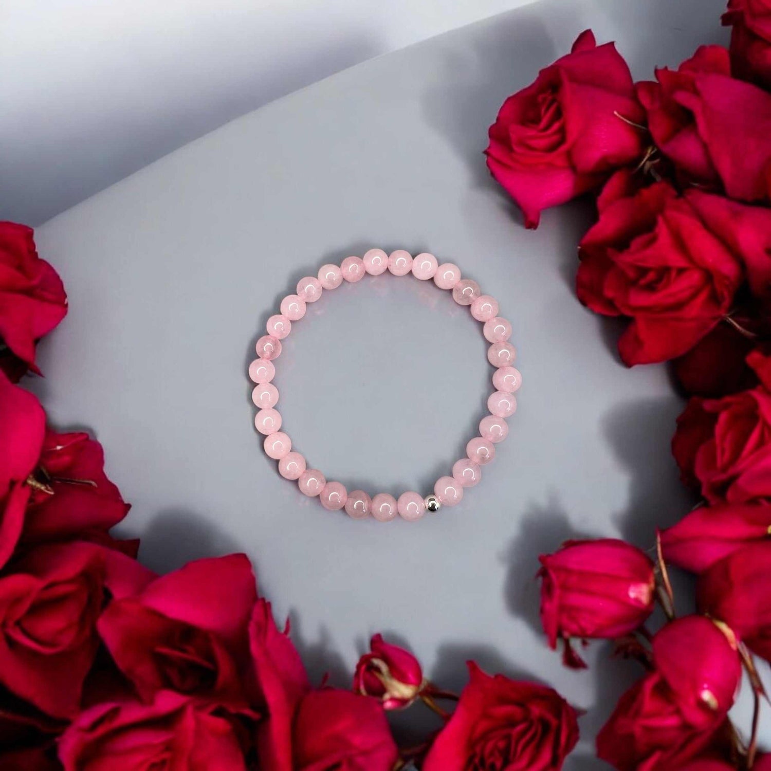 Rosenquarz Armband mit zarten rosa Perlen, handgefertigt, symbolisiert Liebe und Harmonie, präsentiert auf grauem Hintergrund mit roten Rosen.